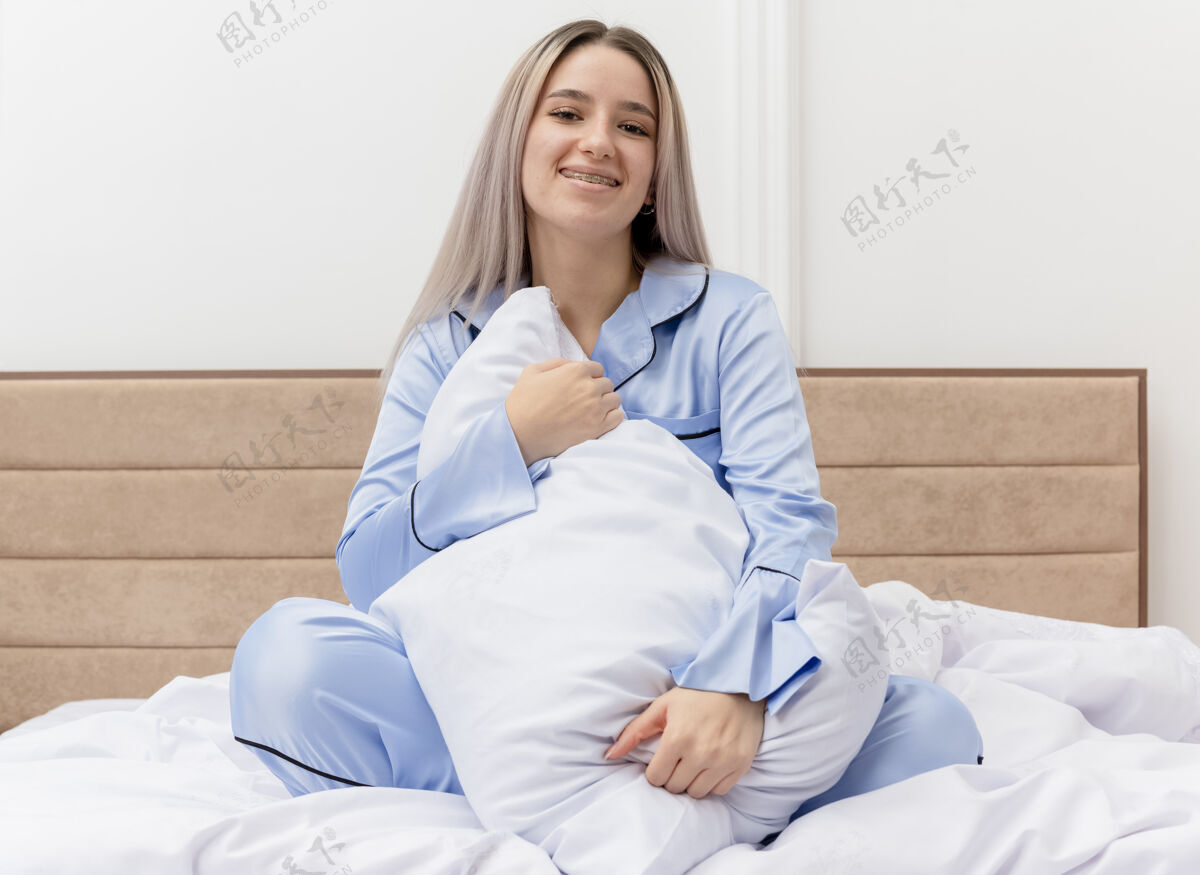脸穿着蓝色睡衣的年轻漂亮女人坐在床上 带着枕头微笑着 幸福的脸躺在卧室里快乐床相机