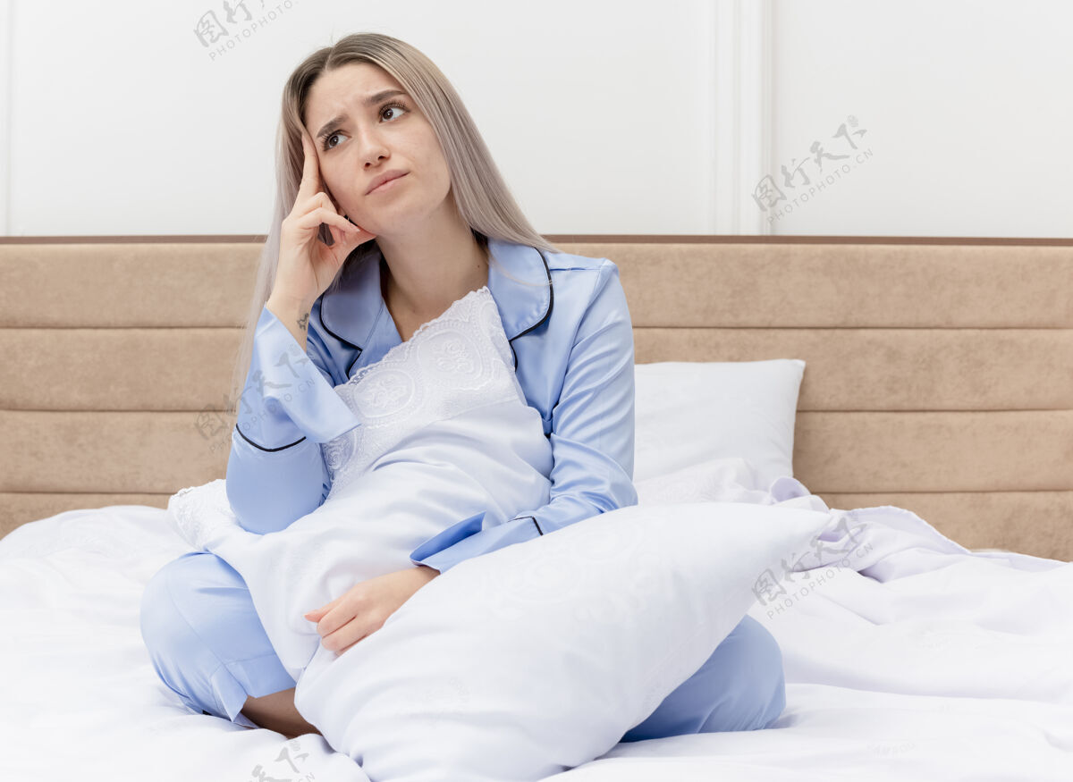 女人穿着蓝色睡衣的年轻美女坐在床上 带着枕头 在卧室里困惑地仰望着蓝色年轻睡衣