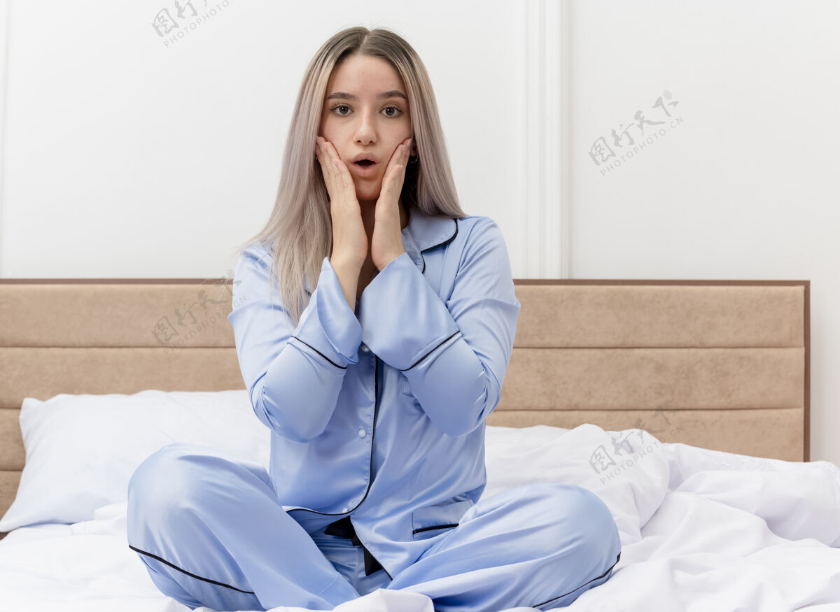 女人穿着蓝色睡衣的年轻漂亮女人坐在床上看着摄像机 在卧室的内部感到惊讶和惊讶蓝色坐着Lookign
