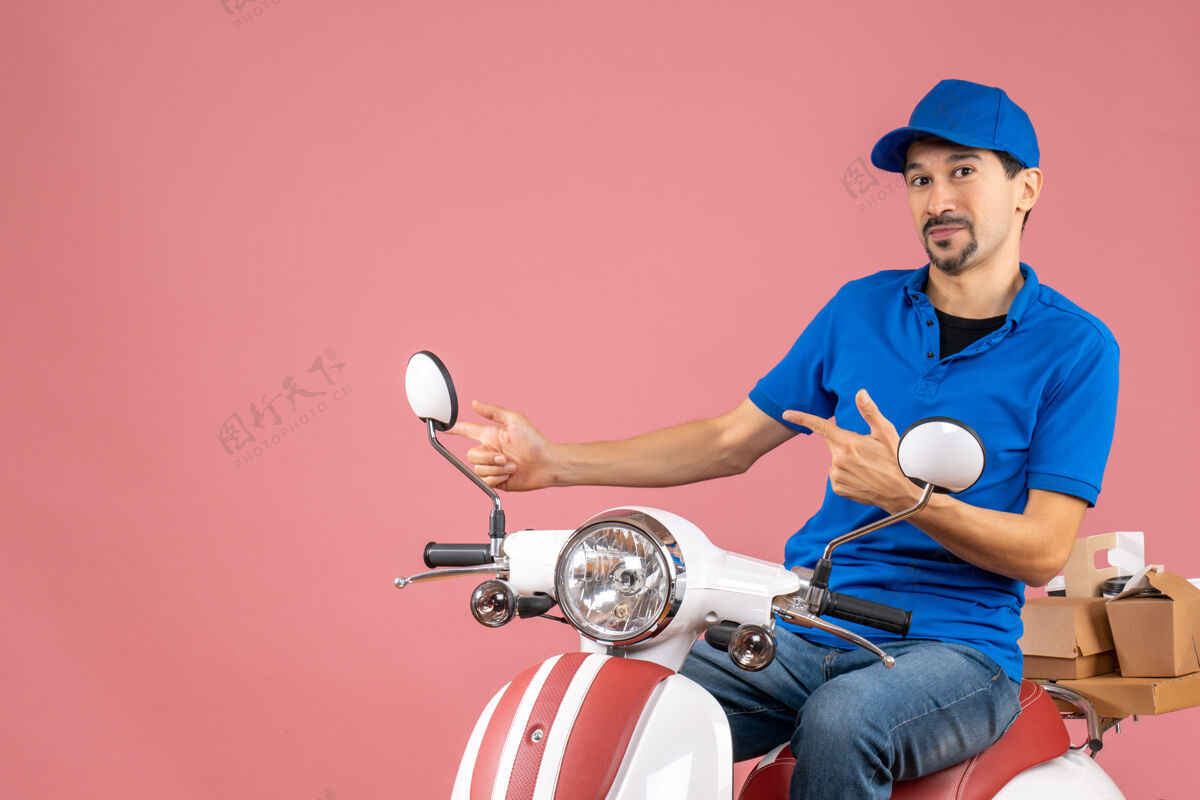足球快递员戴着帽子坐在滑板车上指着粉色背景右侧的东西的正面视图粉彩东西摩托车