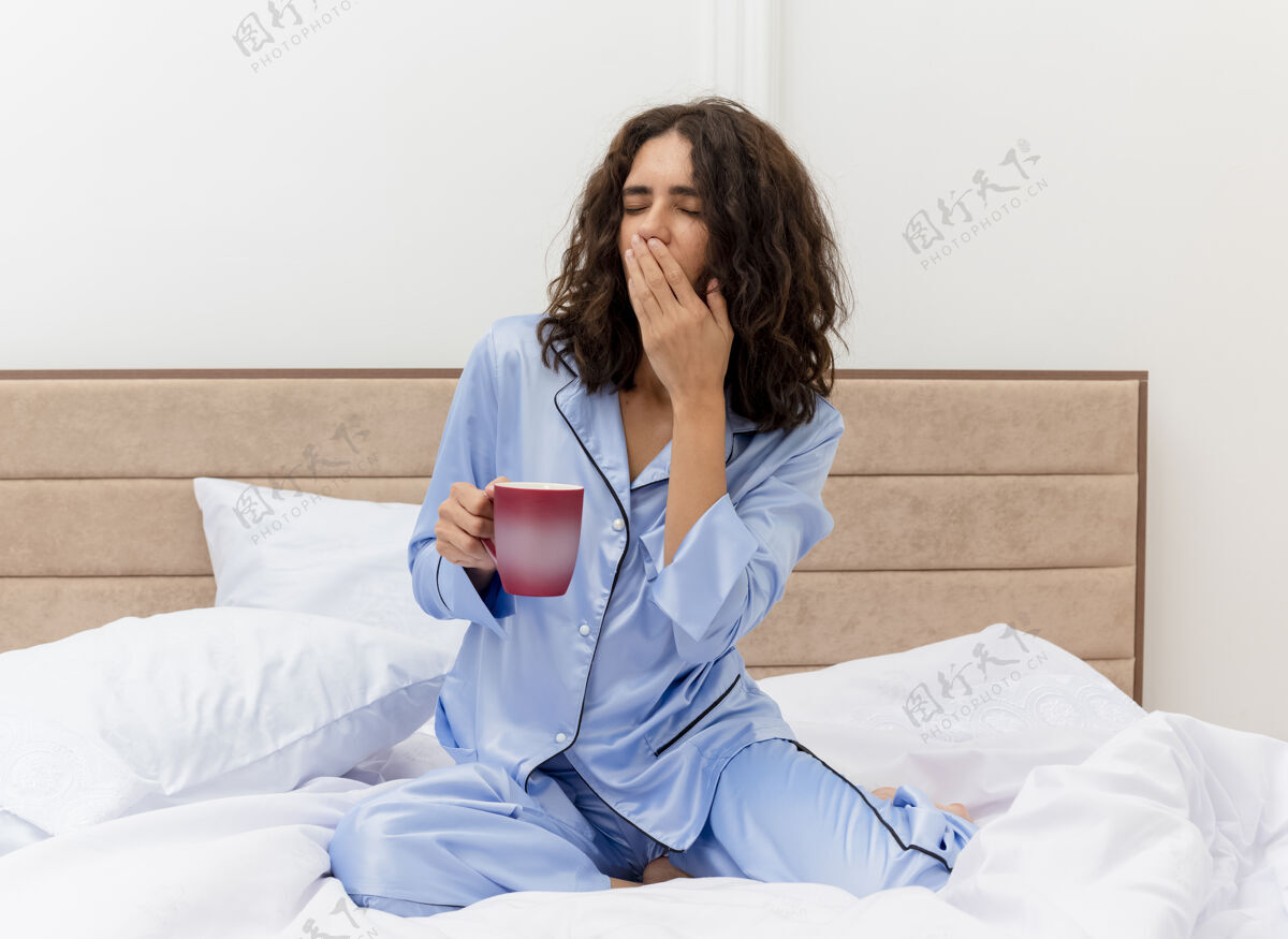 室内穿着蓝色睡衣的有趣年轻漂亮女人坐在床上喝着咖啡醒来打呵欠 感觉卧室里的早晨很疲劳起来醒来杯子