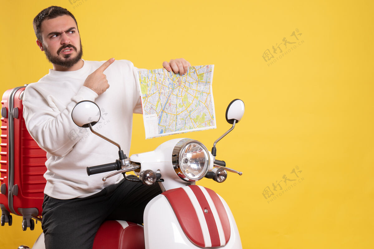 手提箱前视图愤怒的男子坐在摩托车上 手提箱上拿着地图 在孤立的黄色背景上指向上人背景摩托车