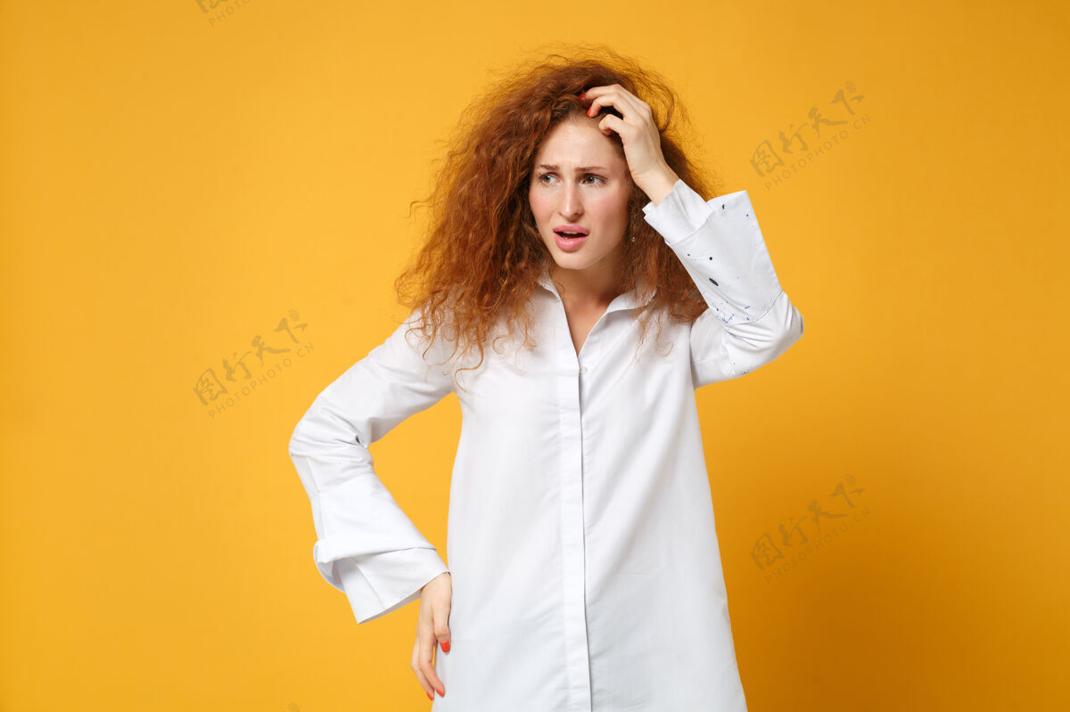 休闲心不在焉的年轻红发女子穿着休闲白衬衫在黄橙色的墙上摆出孤立的姿势橙色女性脸