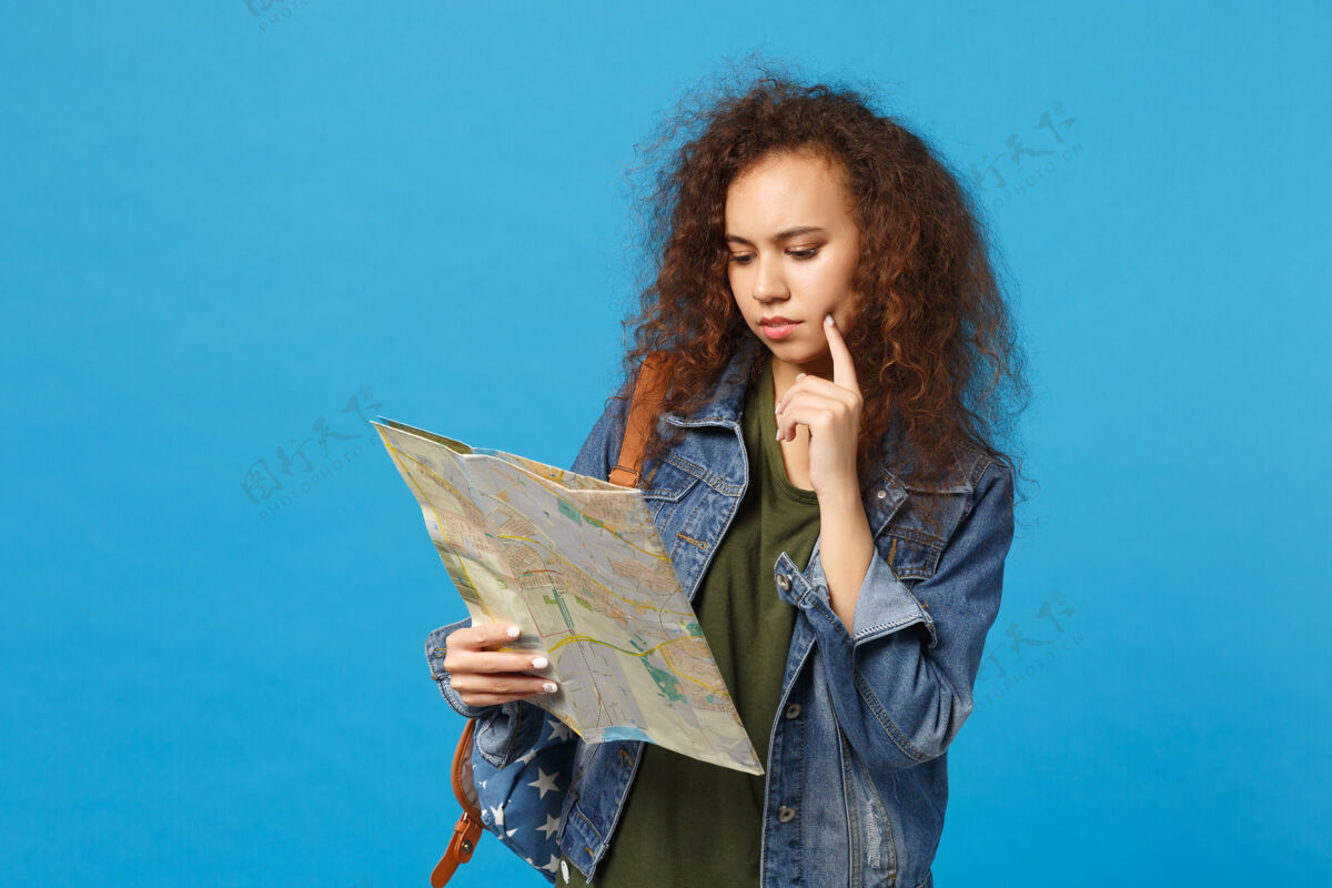学生年轻的非洲裔美国女孩十几岁的学生在牛仔服装 背包举行地图隔离在蓝色的墙上旅游站人