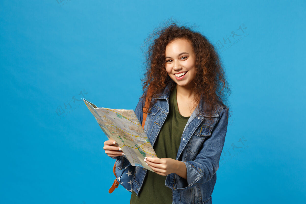旅游年轻的非洲裔美国女孩十几岁的学生在牛仔服装 背包举行地图隔离在蓝色的墙上旅游搜索女人