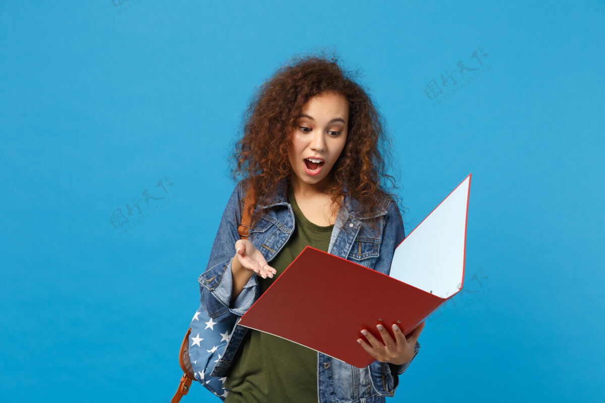 文件年轻的非洲裔美国女孩十几岁的学生在牛仔衣服背包举行文件夹隔离在蓝色墙上非洲红色写作