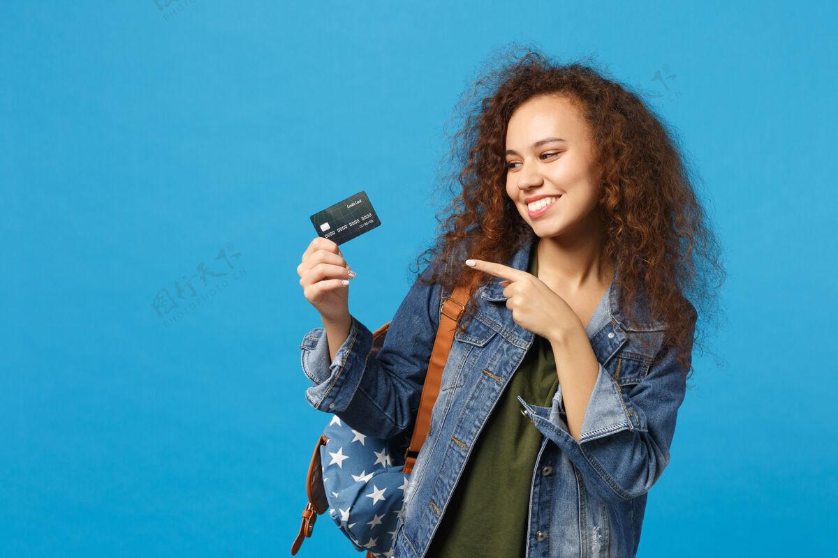 人年轻的非洲裔美国女孩十几岁的学生在牛仔服装 背包举行信用卡隔离在蓝色的墙上青少年购买人