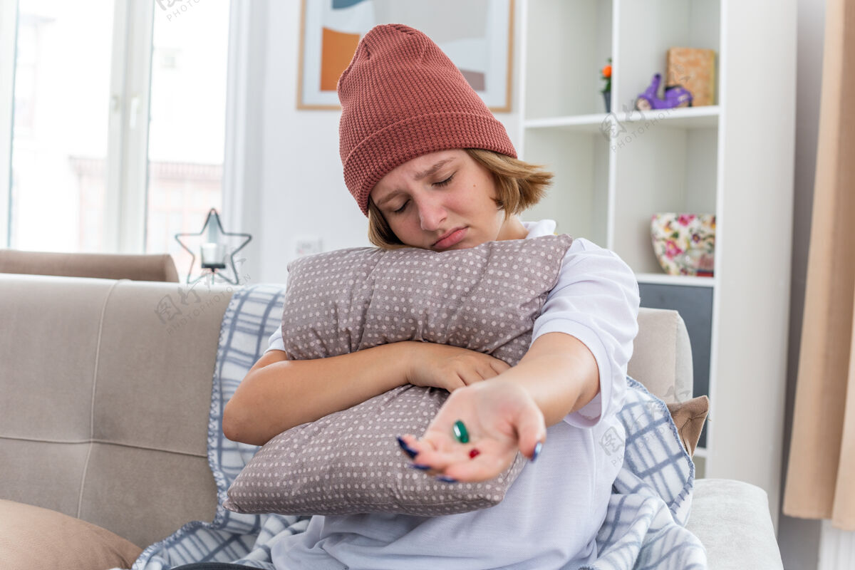 沙发不健康的年轻女子 戴着暖和的帽子 拿着毯子 感觉不舒服 患感冒和流感 抱着枕头和药片 心烦意乱地坐在客厅的沙发上 生病了不适不安帽子