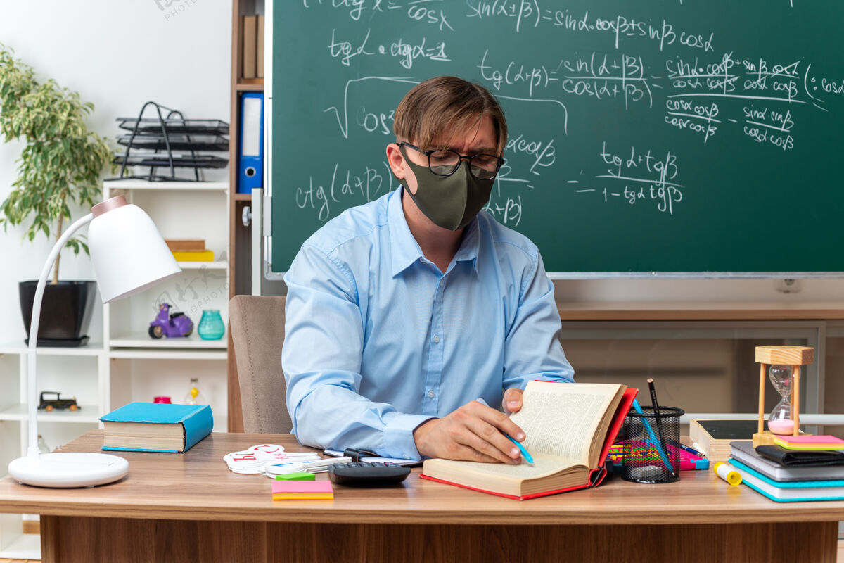 准备年轻的男老师戴着眼镜和面罩备课看起来很自信坐在课桌旁 教室里的黑板前放着书和笔记保护教室年轻