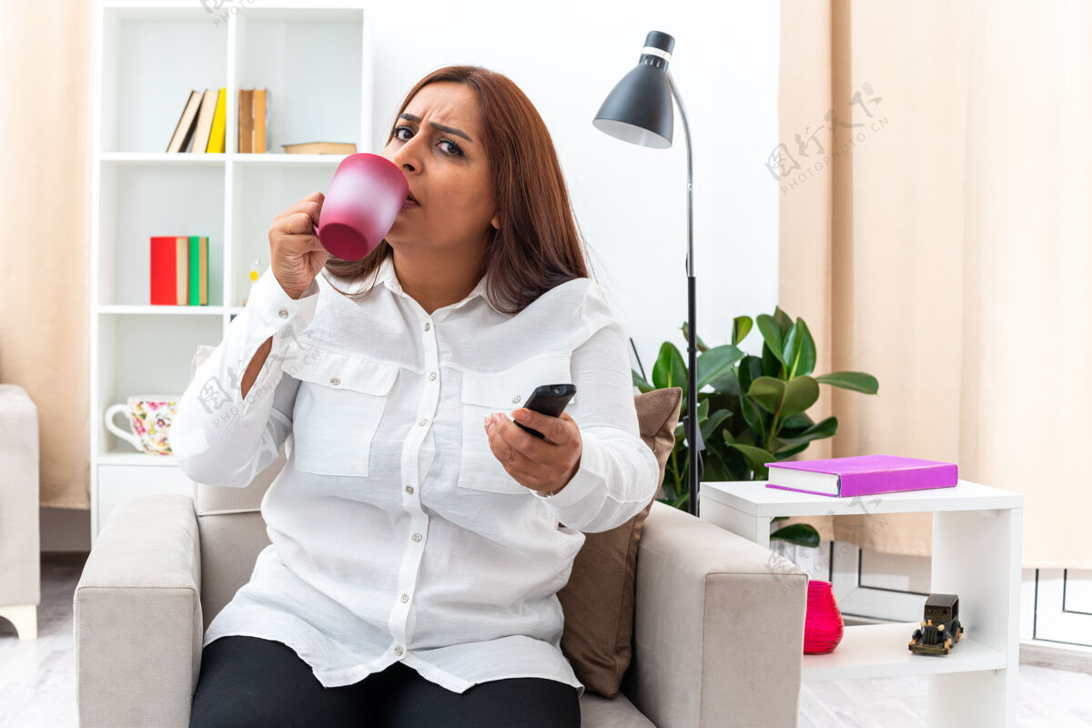 茶穿着白衬衫和黑裤子的女人坐在明亮的客厅里的椅子上看电视喝茶杯杯子光住