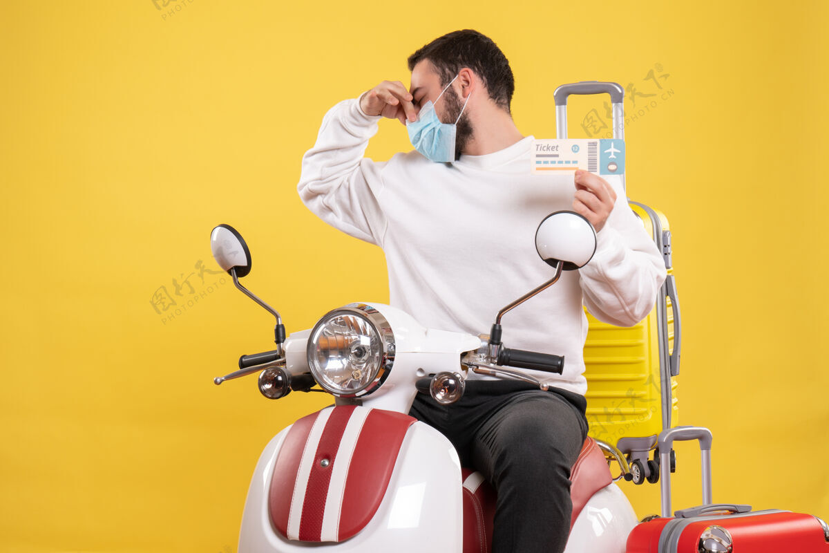 黄色旅行理念俯视图 戴着医用面罩的年轻人坐在摩托车上 黄色手提箱在上面 拿着车票做着难闻的手势球员手提箱手势