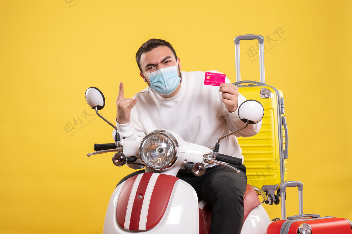 摩托车旅行概念的俯视图 戴着医用面罩的情绪化男人坐在摩托车上 黄色手提箱在上面 手里拿着银行卡黄色银行人