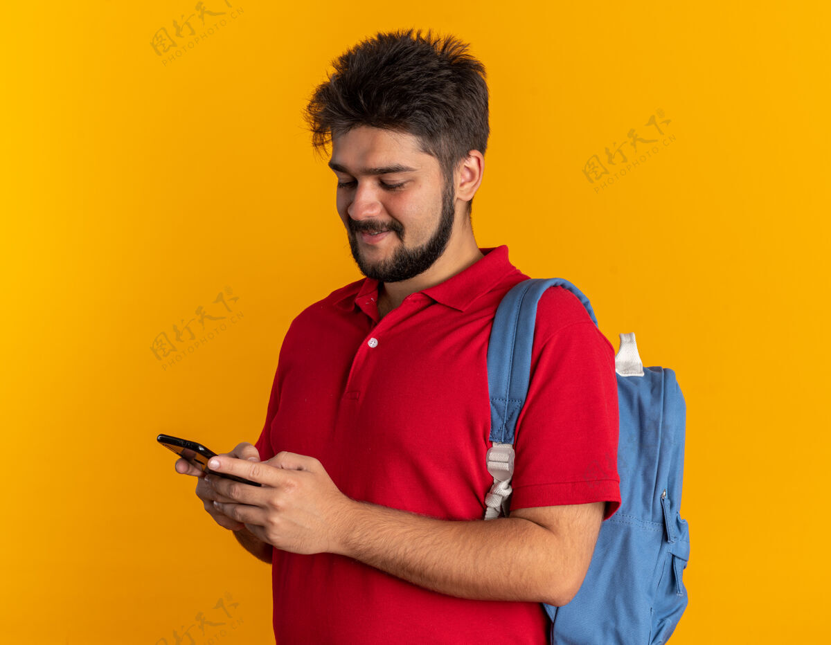 移动年轻的留着胡子的学生 穿着红色马球衫 背着背包 拿着智能手机 面带微笑 站在橙色的墙上 快乐而积极胡须背包手势