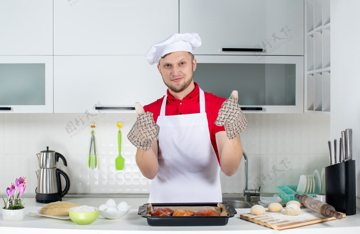 支架俯视图：年轻厨师戴着支架站在桌子后面 上面放着糕点和鸡蛋磨碎器 在白色厨房里做着“ok”的手势手势顶部磨碎器