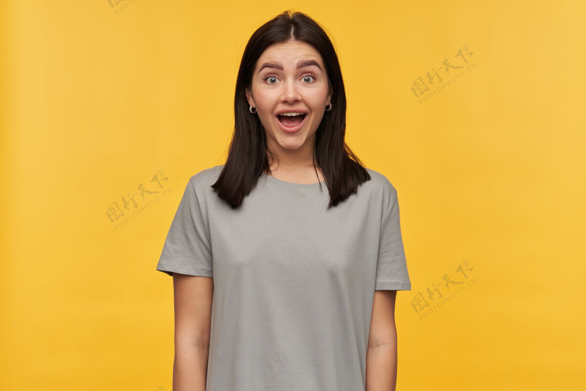 欢呼一头黑发 张嘴 身穿灰色T恤的年轻女子在黄色的墙上显得很兴奋女士开放情绪