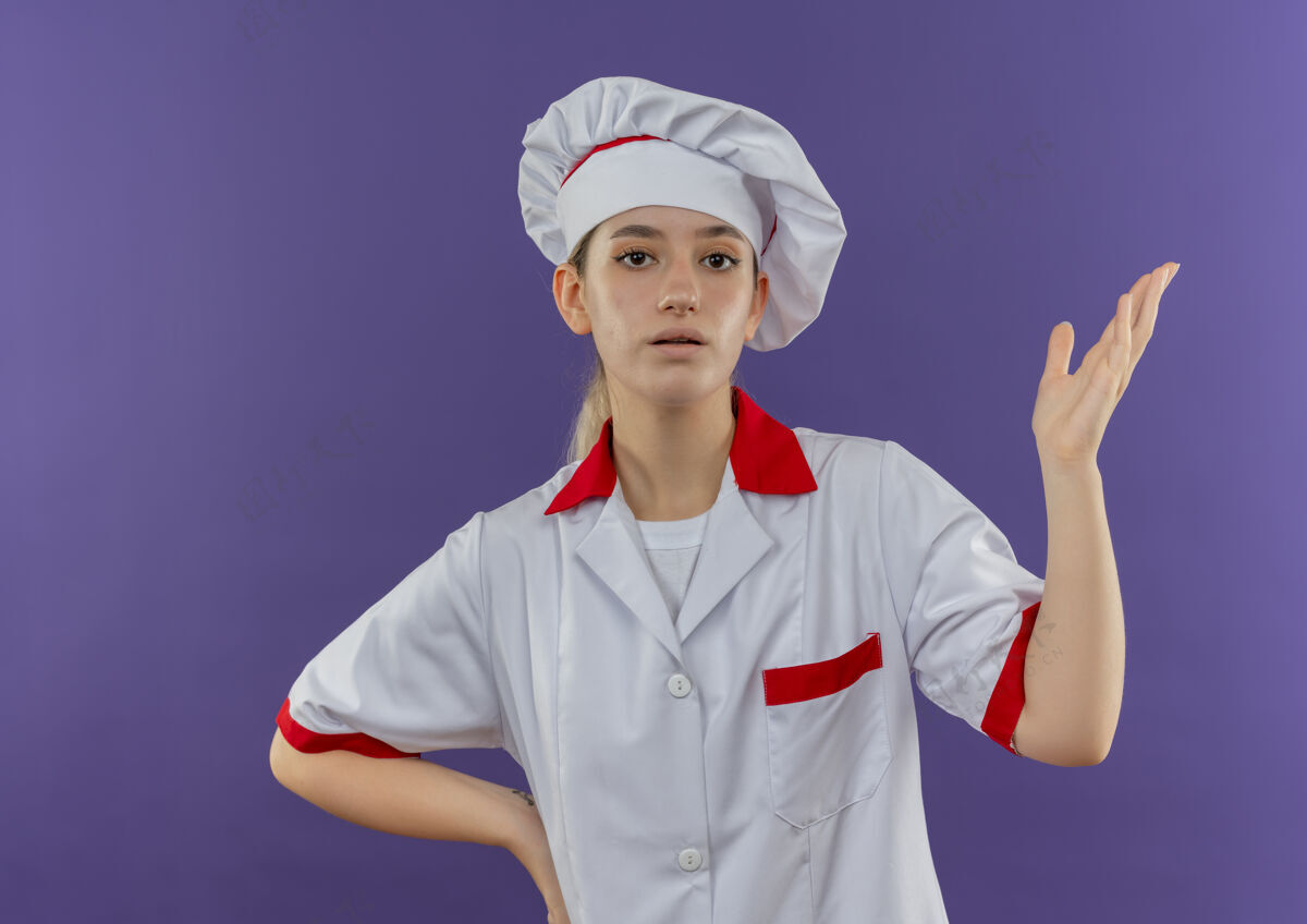 印象穿着厨师制服的年轻漂亮厨师举起手来 把另一只手放在紫色墙壁上的腰部举另一个年轻