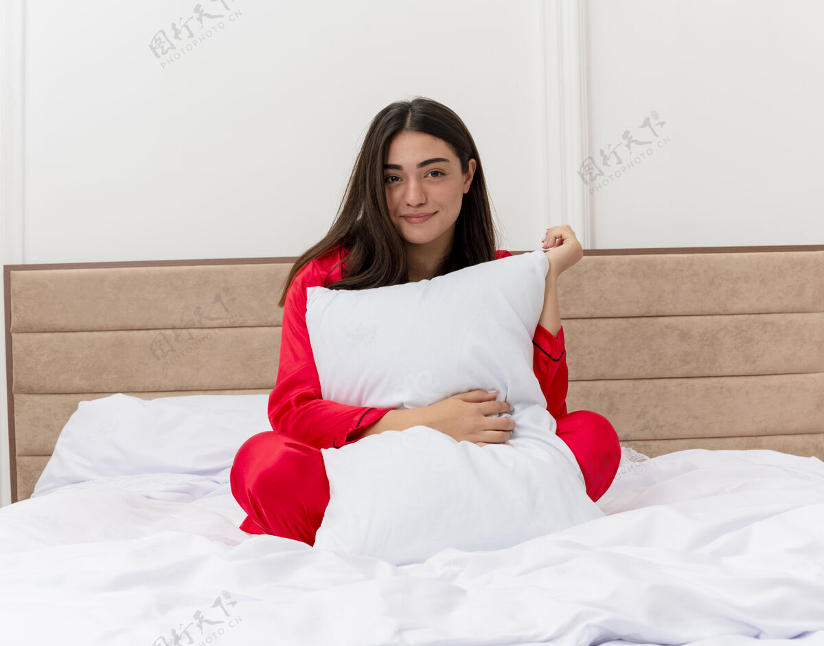 漂亮穿着红色睡衣的年轻漂亮女人坐在床上 枕着枕头 在卧室里笑容可掬室内女士快乐