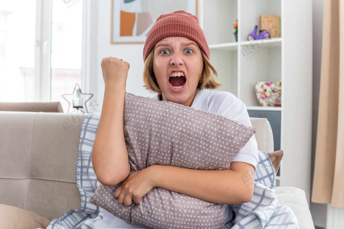表情愤怒的不健康的年轻女子 戴着暖和的帽子 拿着毯子 看起来不舒服 患感冒和流感 抱着枕头 握紧拳头 带着咄咄逼人的表情 坐在客厅的沙发上流感生气年轻
