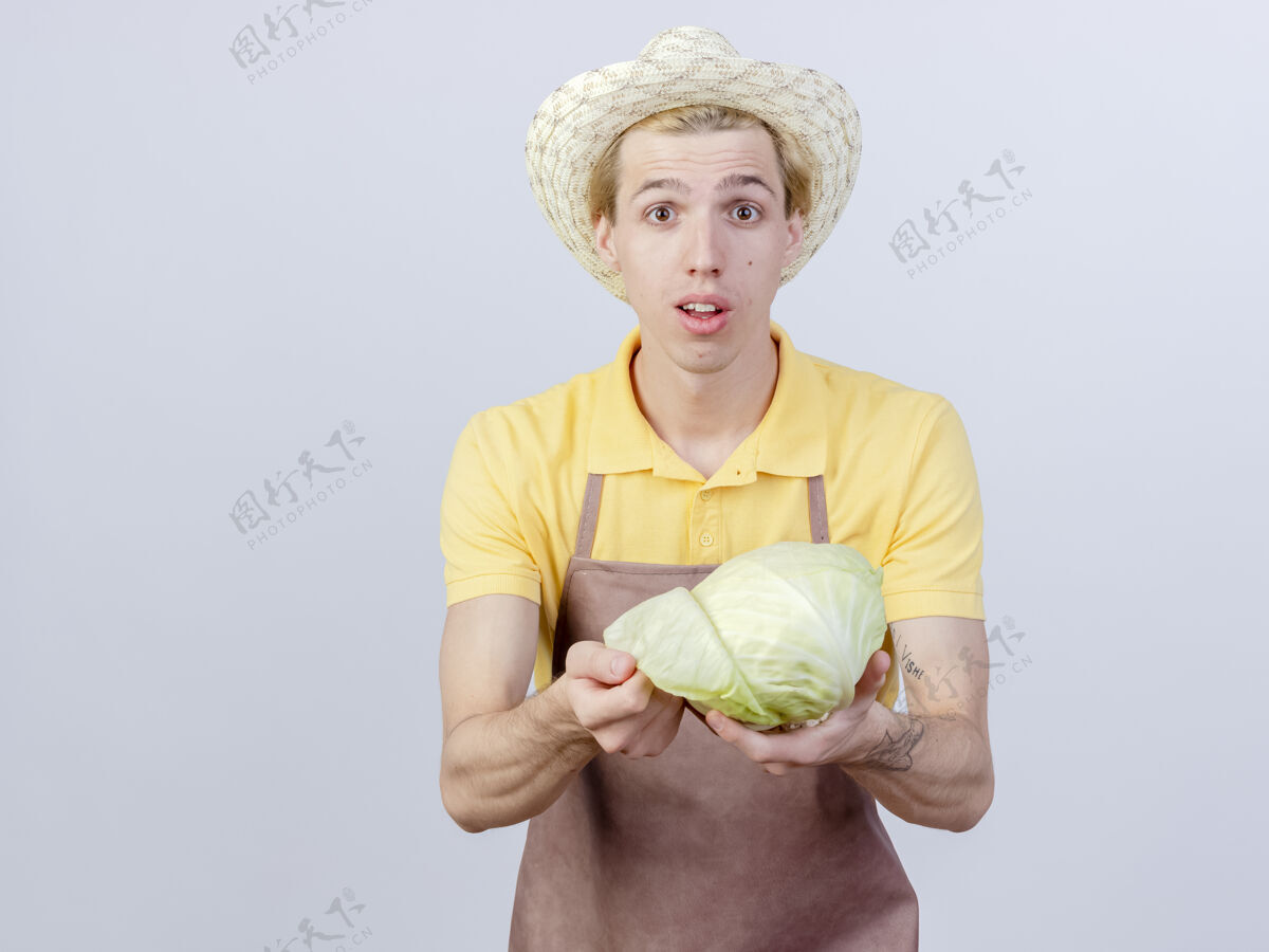 男人年轻的园丁 穿着连体衣 戴着帽子 手里拿着白菜 脸上带着微笑微笑帽子站着