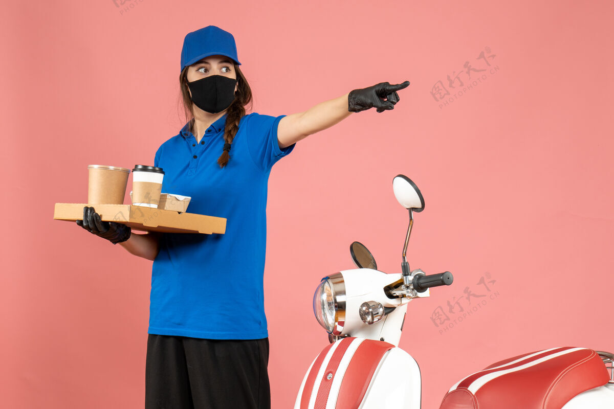 粉彩俯视图：戴着医用口罩手套的快递员女孩站在摩托车旁 手里拿着咖啡小蛋糕 在柔和的桃色背景上指向前方桃子向前咖啡