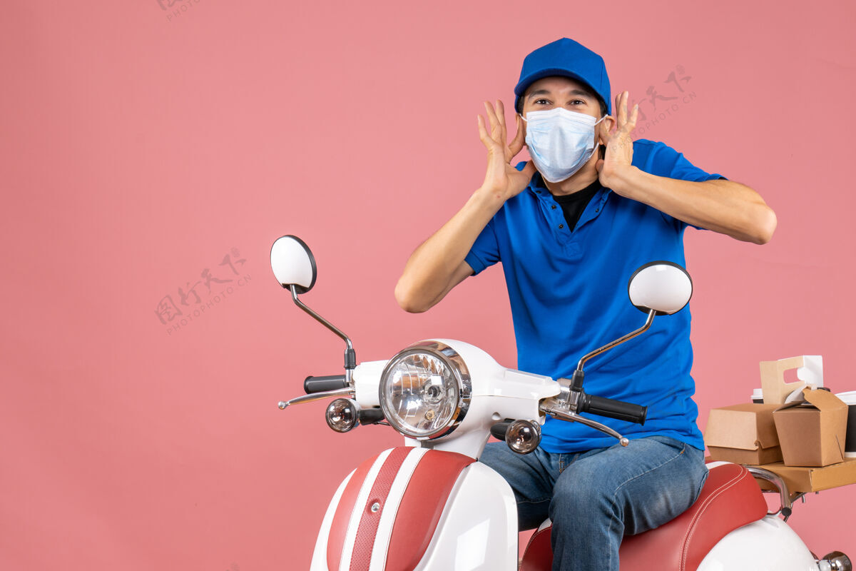 滑板车一个戴着医用面罩 戴着帽子 坐在粉彩桃色背景下的滑板车上的震惊的送货员的正视图医疗摩托车送货员