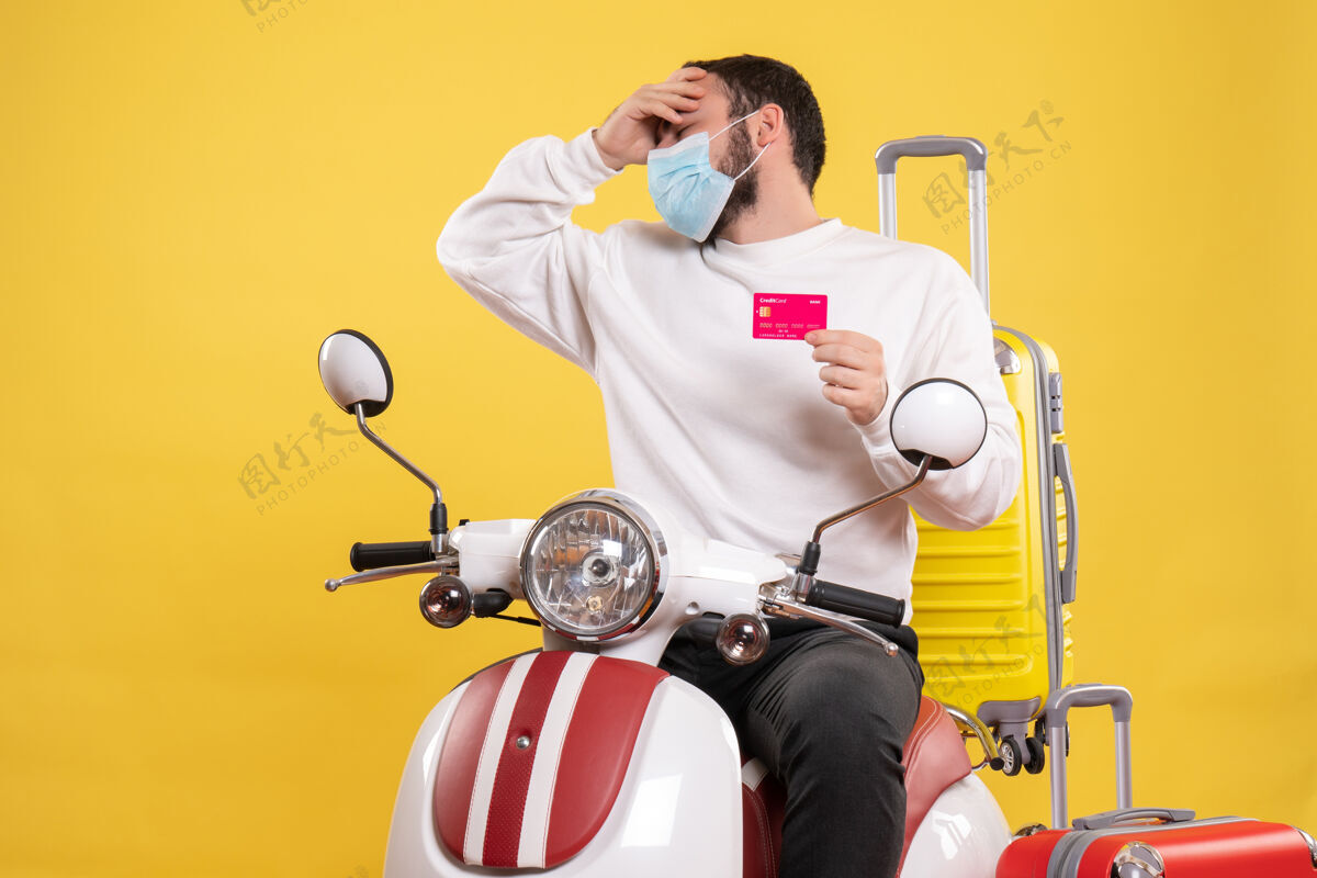 年轻人旅行概念的正面图 戴着医用面罩的问题青年坐在摩托车上 黄色手提箱 手里拿着银行卡微笑面具成人