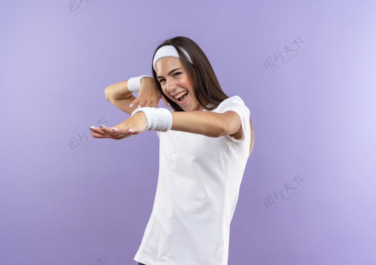 头带快乐漂亮的运动女孩戴着头带和腕带伸出手与另一个靠近头部隔离在紫色墙上欢乐另一个女孩