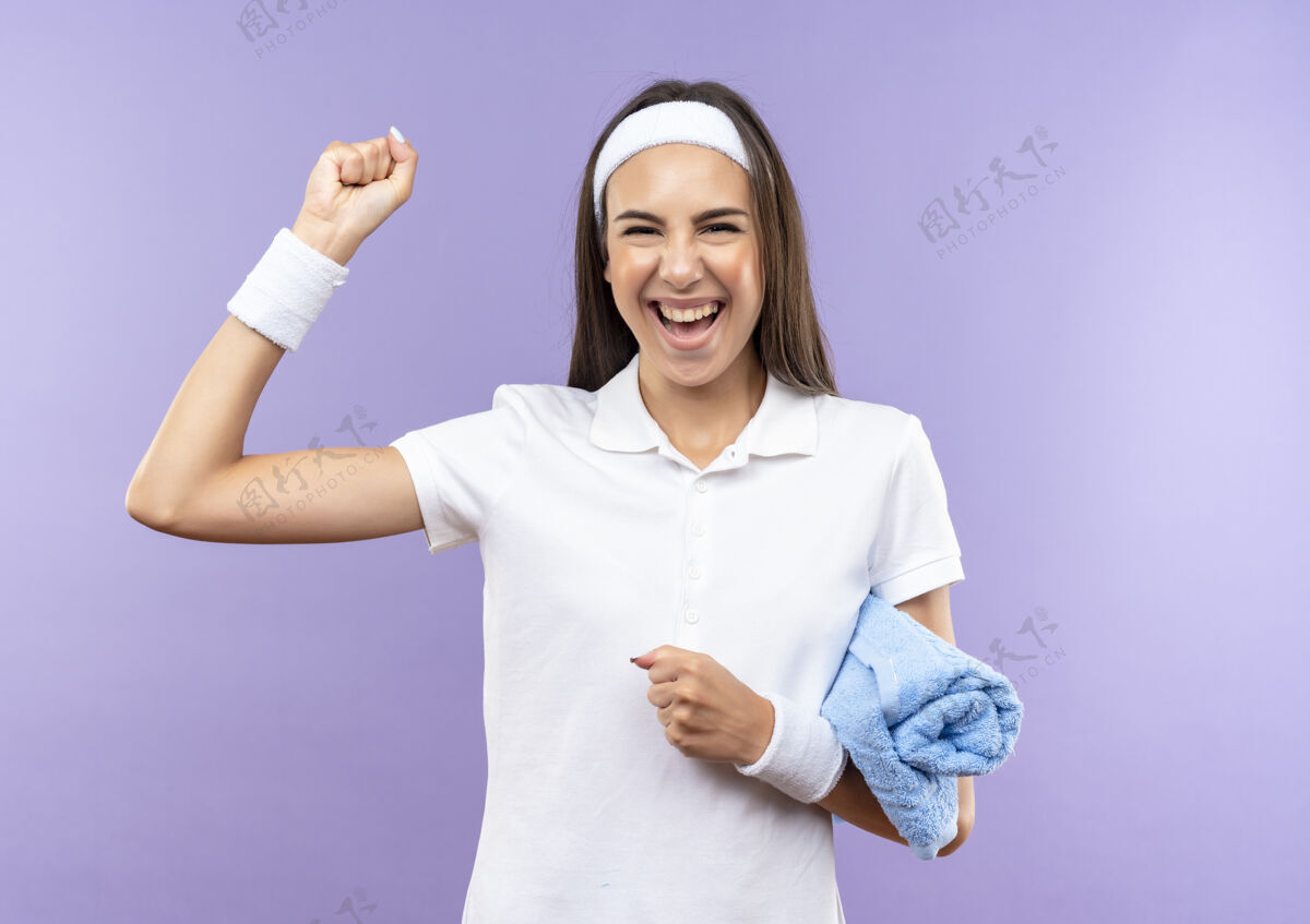 拳头快乐漂亮的运动女孩戴着头带和手环拿着毛巾 举起拳头孤立在紫色的墙上腕带提高抱着