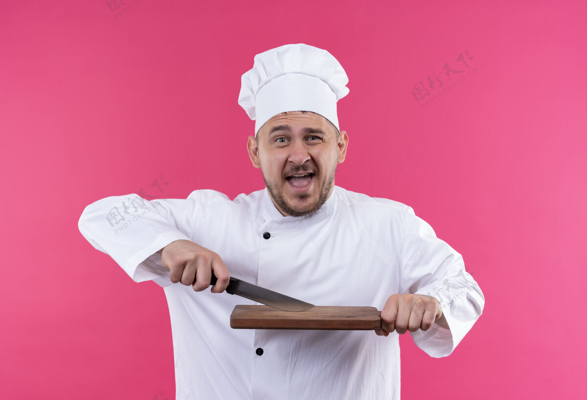 制服快乐的年轻帅哥厨师穿着厨师制服拿着刀和砧板隔离在粉红色的墙上烹饪刀持有