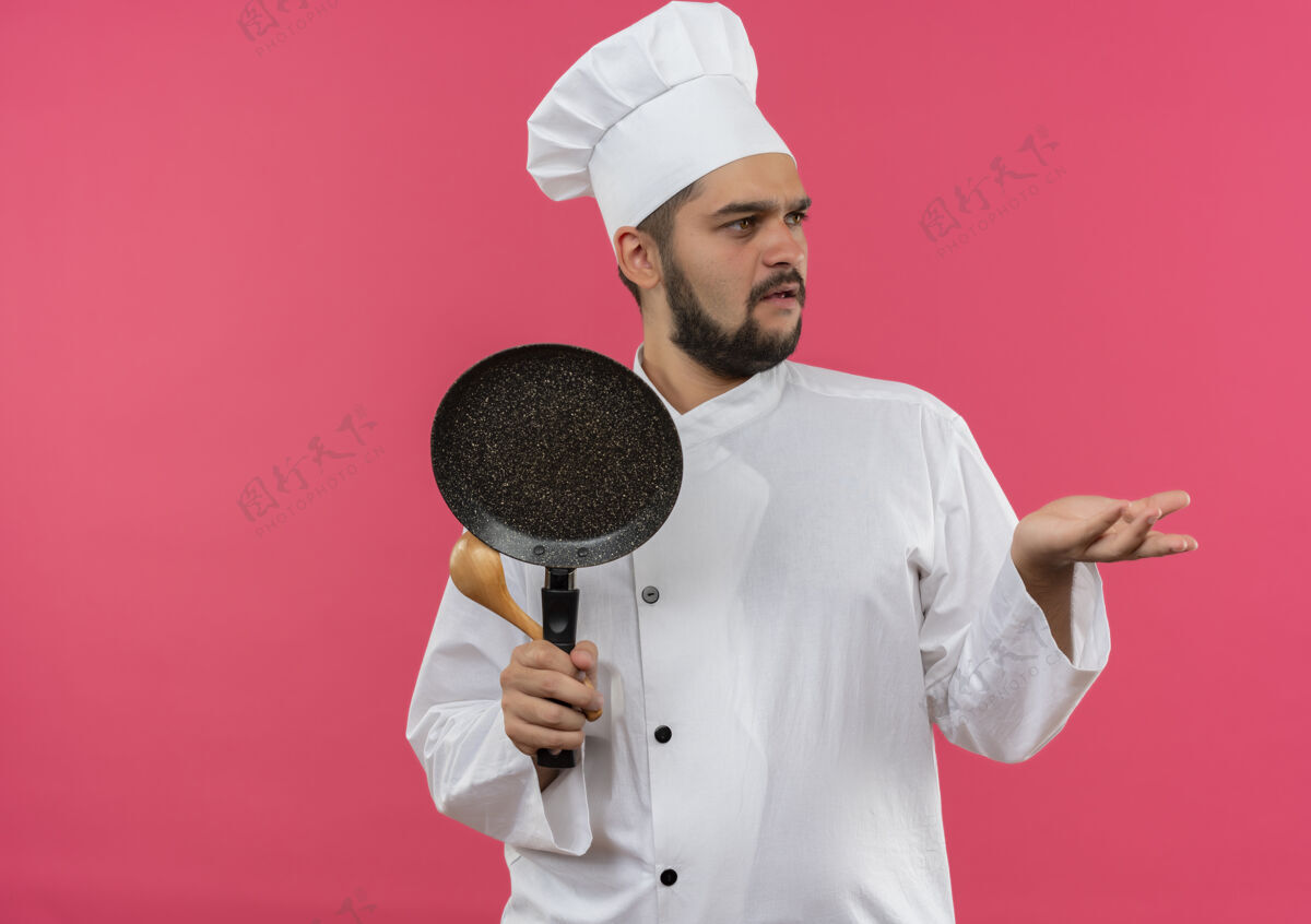 年轻困惑的年轻男厨师穿着厨师制服 拿着煎锅和勺子 看着侧面 在粉色的墙壁上显示出一只空的手 还有复制空间困惑手烹饪