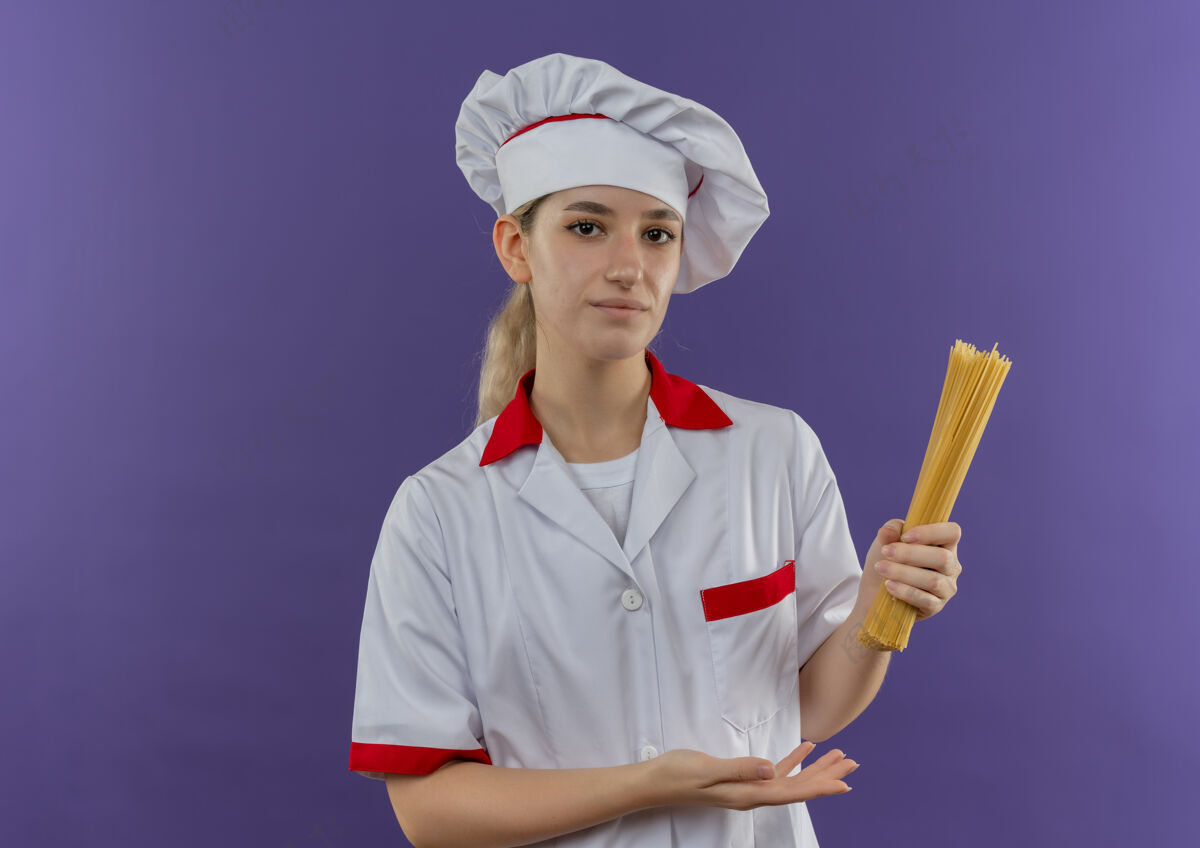 意大利面身着厨师制服的自信年轻漂亮的厨师手拿着并指着紫色墙上孤立的意大利面年轻厨师烹饪