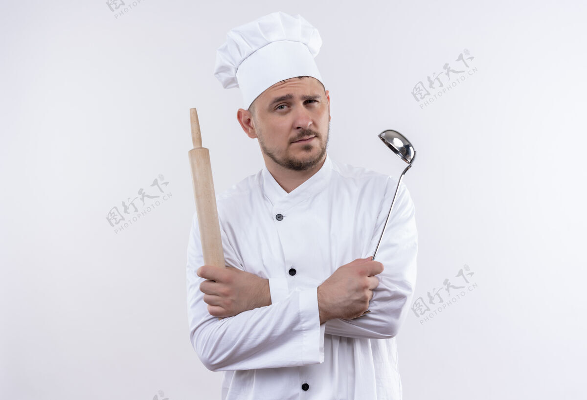 卷自信的年轻帅哥厨师身着厨师制服 拿着勺子和擀面杖 站在隔离的白色墙壁上 留着复制空间别针自信厨师