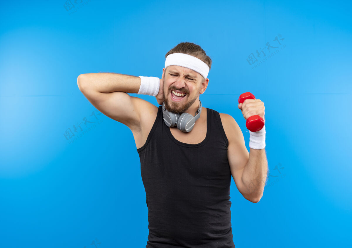 腕带疼痛的年轻英俊的运动型男子戴着头带和腕带 戴着耳机 脖子上抱着哑铃 手放在脖子上 隔离在蓝色的墙上疼围着哑铃