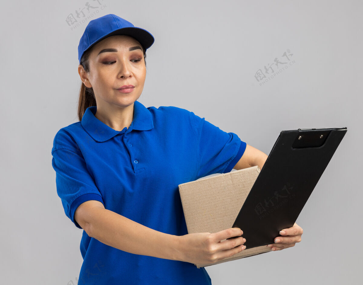 女人身穿蓝色制服 头戴鸭舌帽 拿着纸板箱和剪贴板的年轻女送货员站在白色的墙上 看上去很自信剪贴板交货举行