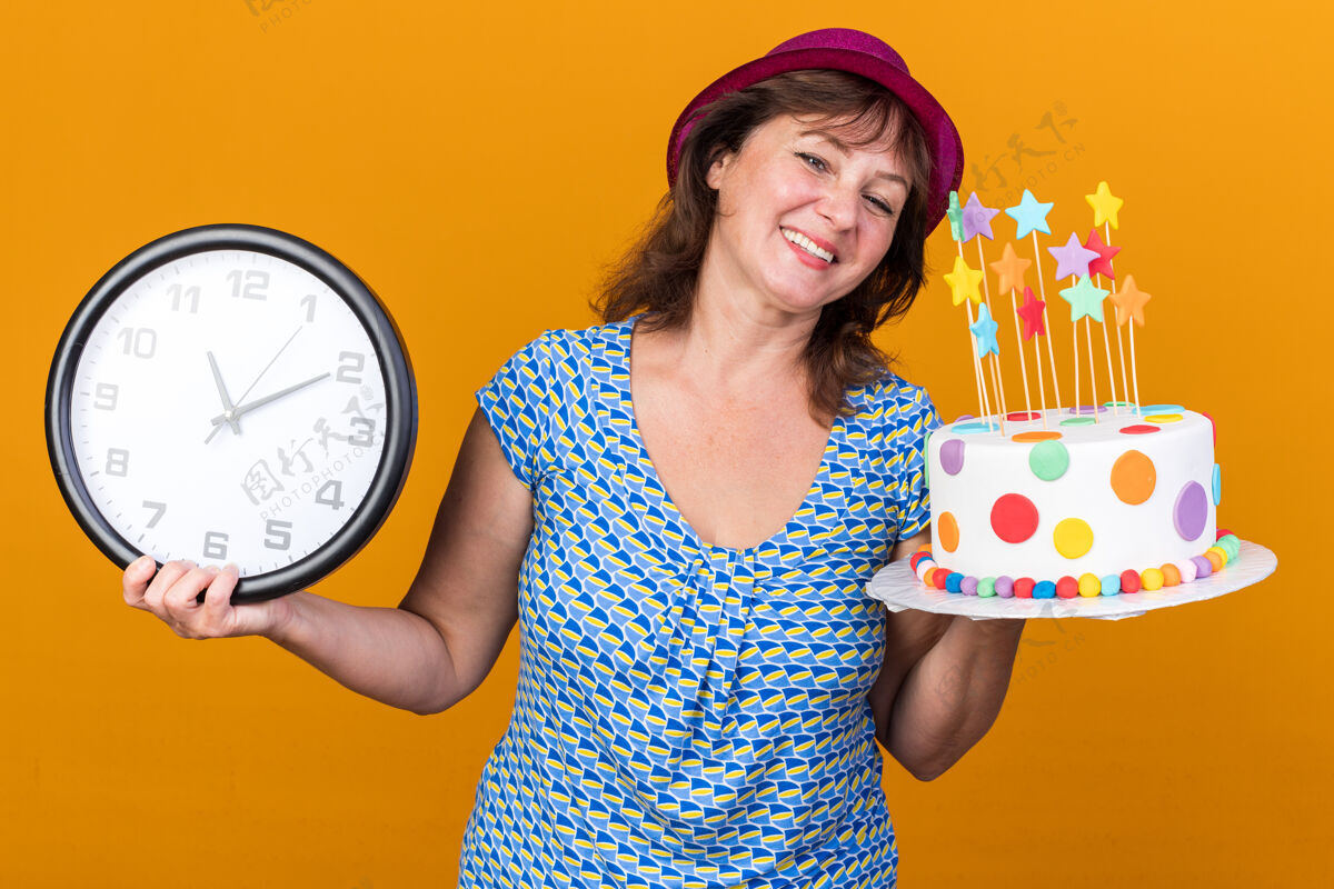 生日快乐快乐的中年妇女 戴着派对帽 拿着生日蛋糕和挂钟 站在橘色的墙上欢笑着庆祝生日派对中间举行欢呼