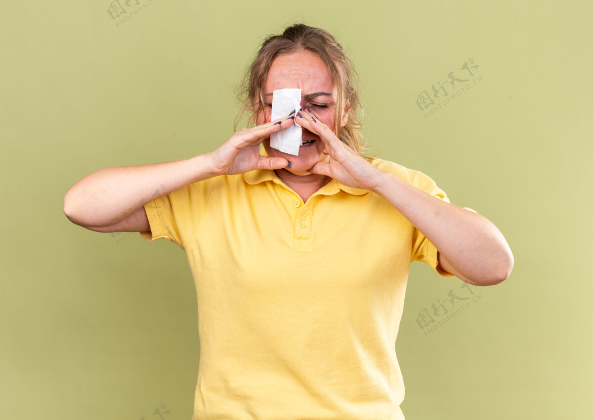 头痛穿着黄衬衫的不健康的女人站在绿墙上流鼻涕 头痛得厉害 感觉很可怕感情女人鼻子