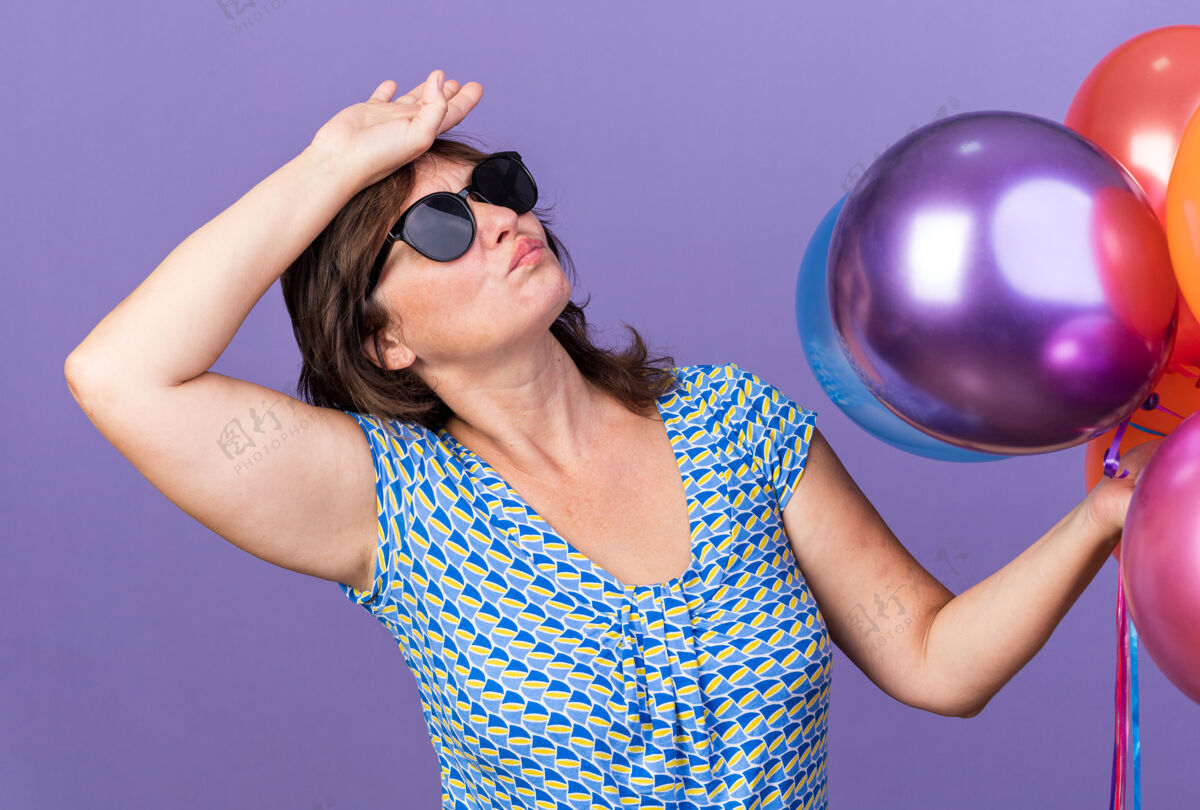 累中年妇女戴着眼镜 手里拿着一堆五颜六色的气球 抬头看着疲惫无聊的手放在头上 站在紫色的墙上庆祝生日派对手眼镜五颜六色