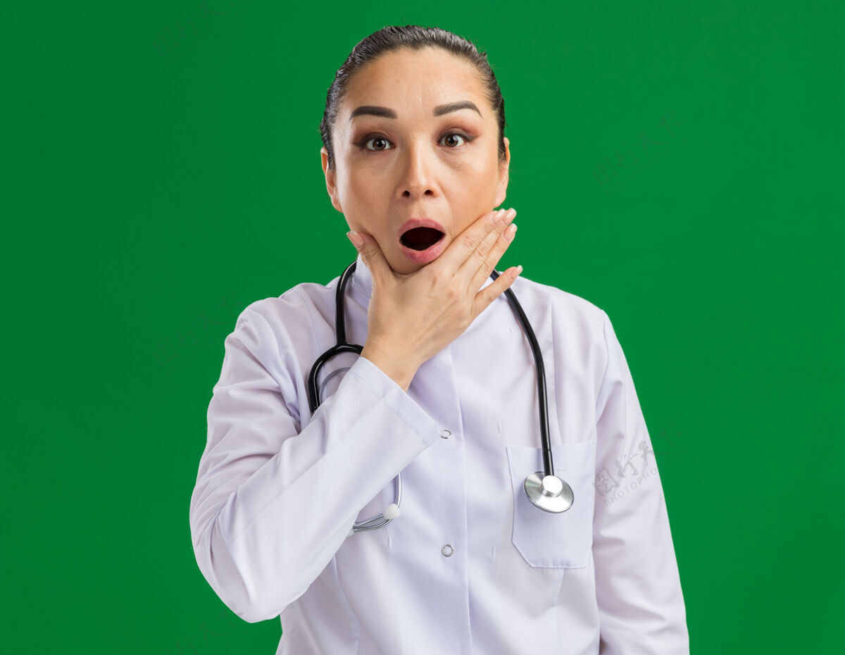 惊喜身穿白色药衣 脖子上戴着听诊器的年轻女医生站在绿色的墙壁上 既惊讶又惊讶医生药物外套
