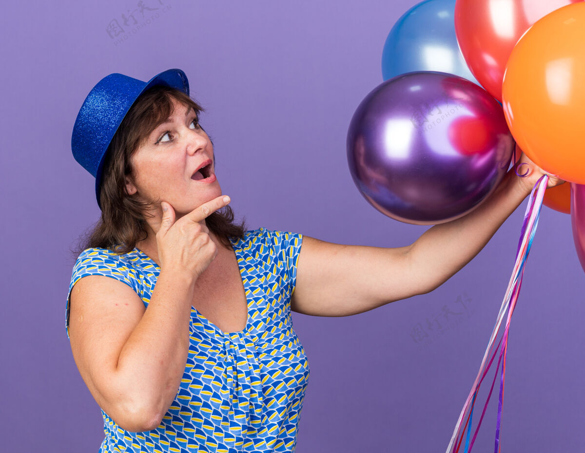 年龄中年妇女戴着派对帽 手里拿着一堆五颜六色的气球 站在紫色的墙上惊讶地看着他们庆祝生日五颜六色惊人女人