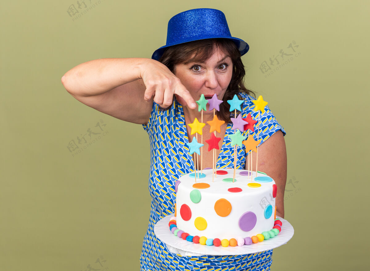 指向中年妇女戴着派对帽 拿着生日蛋糕 用食指指着它 开心又惊喜地站在绿色的墙上庆祝生日派对手指举行生日