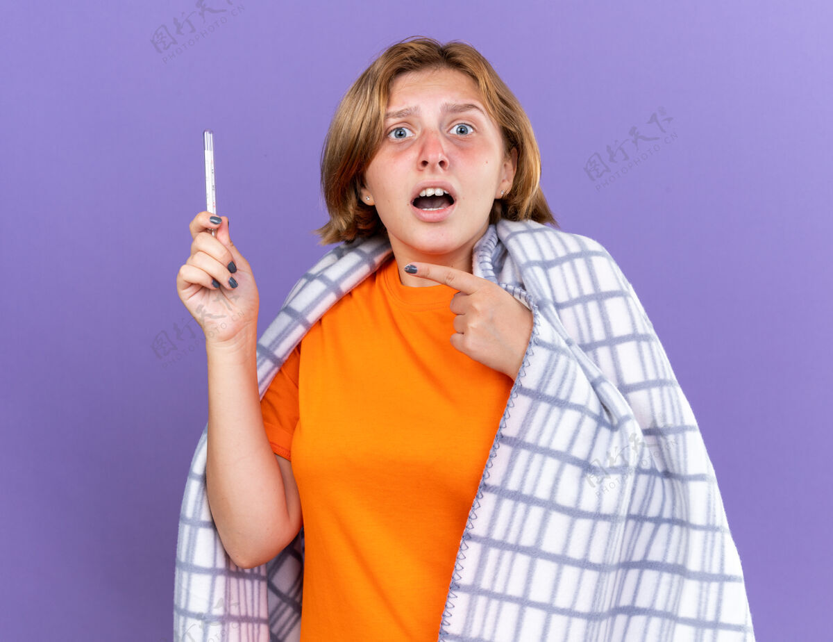 使用不健康的年轻女子裹着温暖的毯子 感觉不舒服 患流感 发烧 用温度计测量体温 站在紫色的墙上 看着它 很担心女人暖和年轻