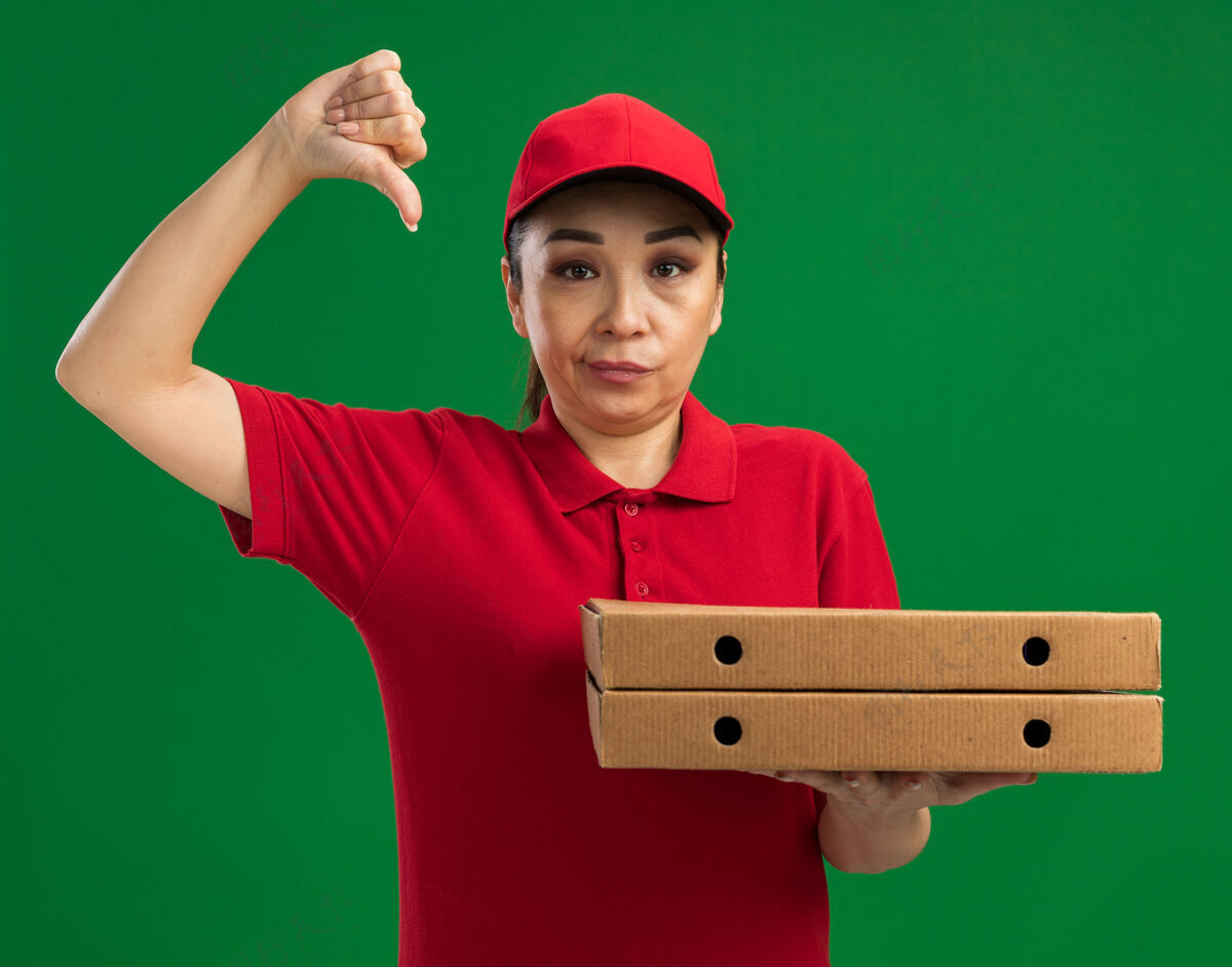 帽子身穿红色制服 头戴鸭舌帽 拿着比萨饼盒的年轻送货员不高兴地竖起大拇指站在绿色的墙上拇指年轻人盒子