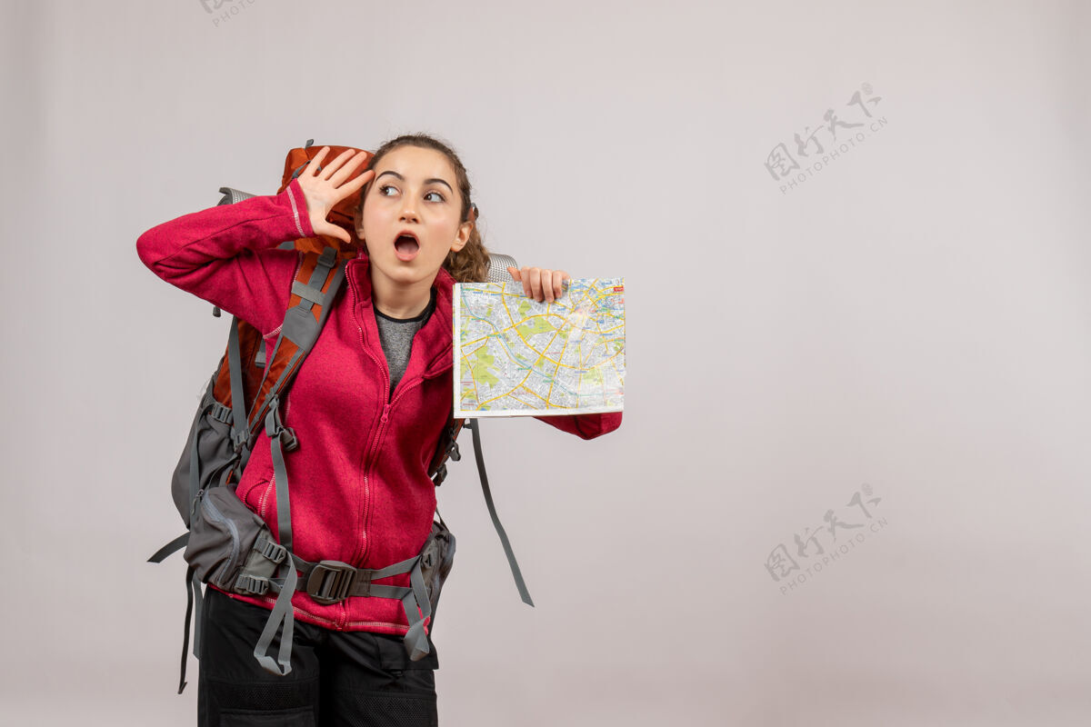 时尚带着大背包的年轻旅行者惊讶地把地图举在灰色的地图上地图成人背包