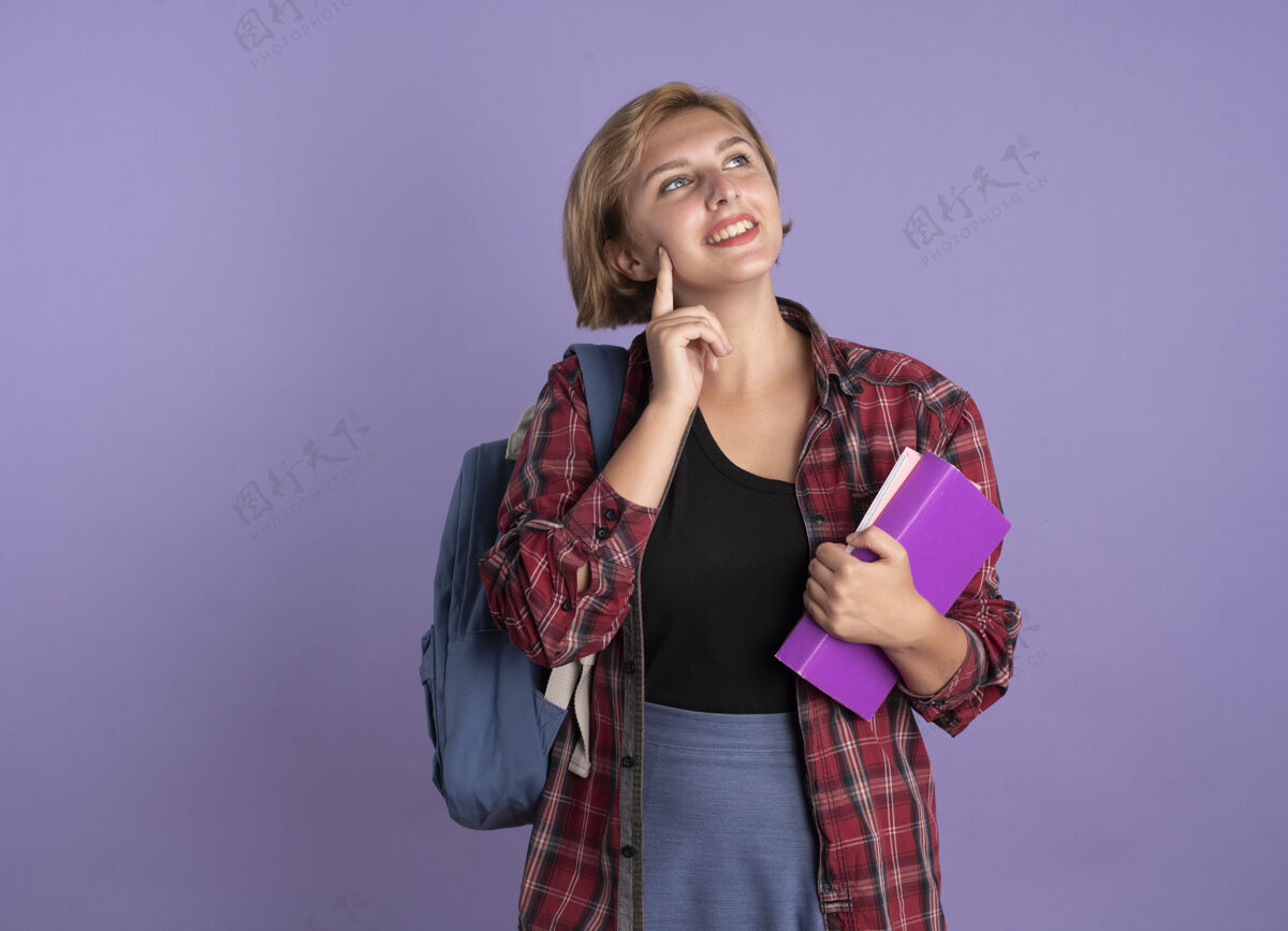 拿着微笑着的年轻斯拉夫学生女孩背着背包把手指放在脸上拿着书和笔记本抬头看学生斯拉夫背包