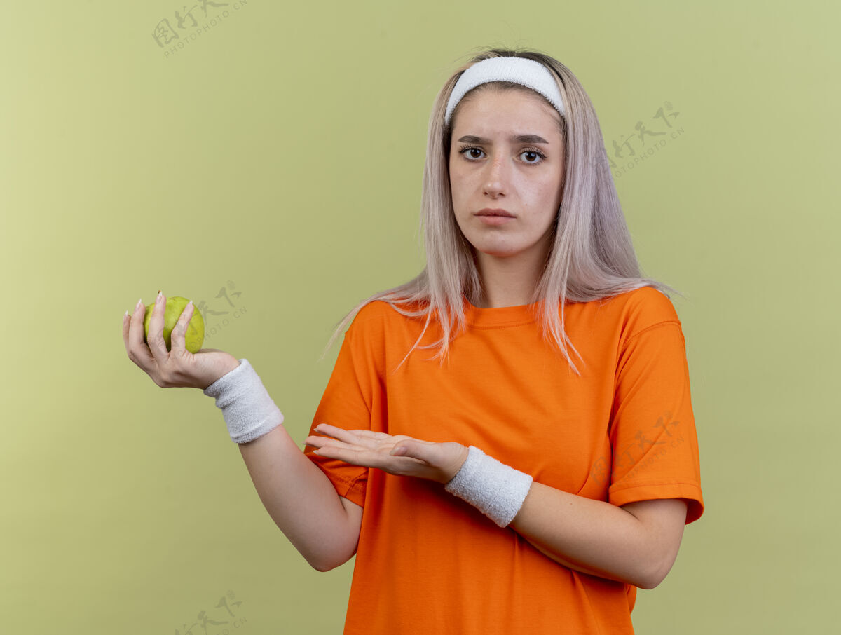 背带无知的年轻白人运动女孩戴着背带 戴着头带和腕带 拿着苹果 指着苹果女孩点举行