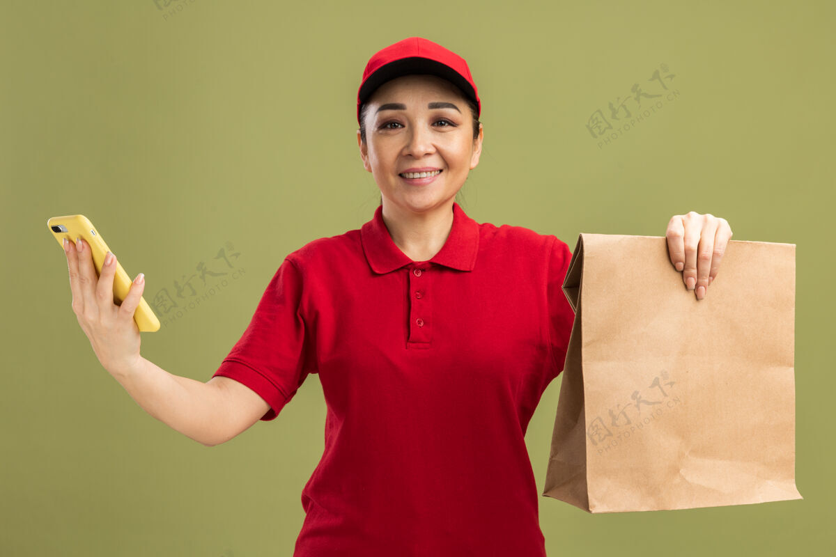 微笑身着红色制服 戴着帽子 手持纸包和智能手机 脸上带着微笑的年轻送货员站在绿色的墙上帽子持有脸