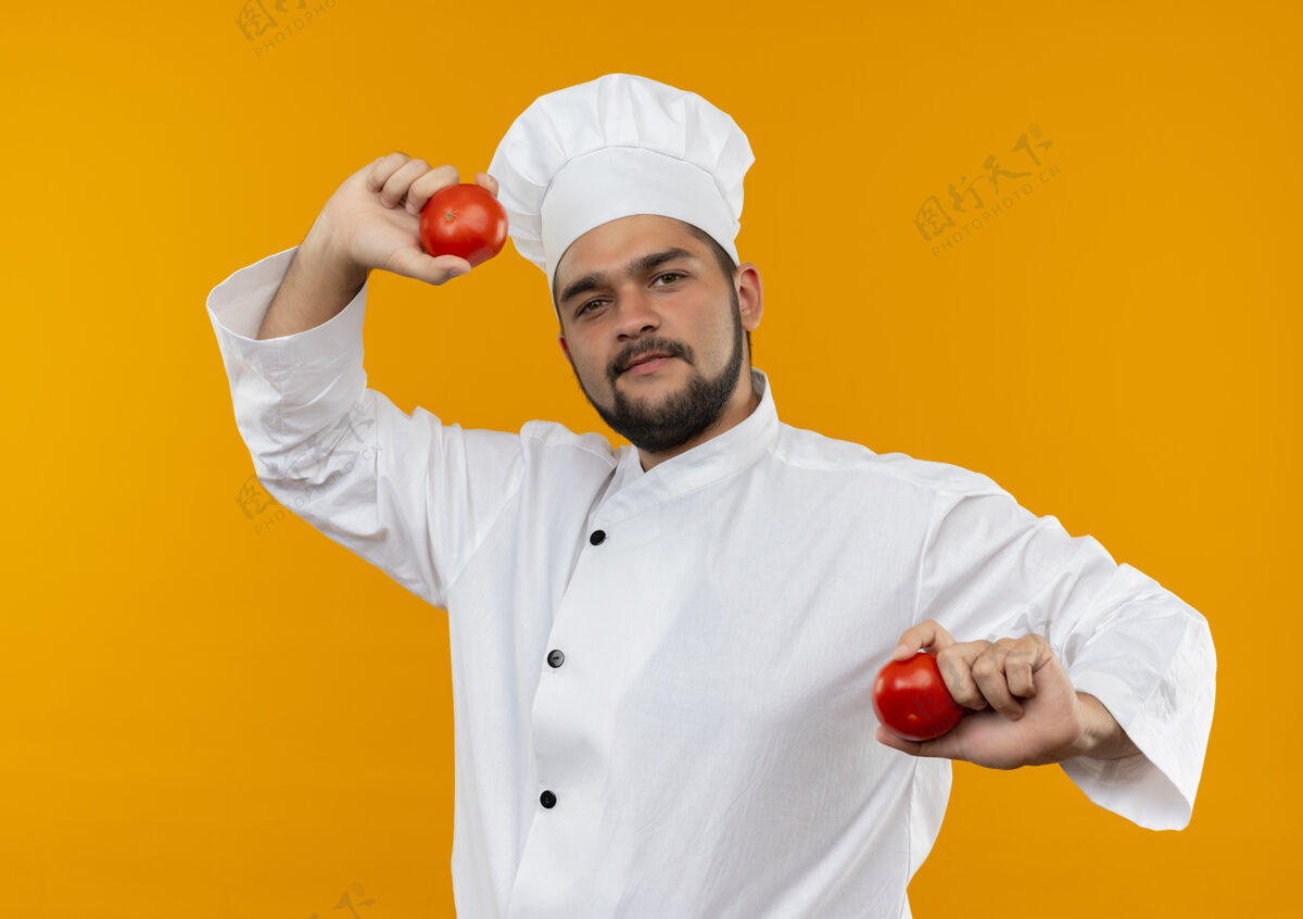 番茄自信的年轻男性厨师身着厨师制服 手里拿着隔离在橙色墙上的西红柿年轻制服厨师