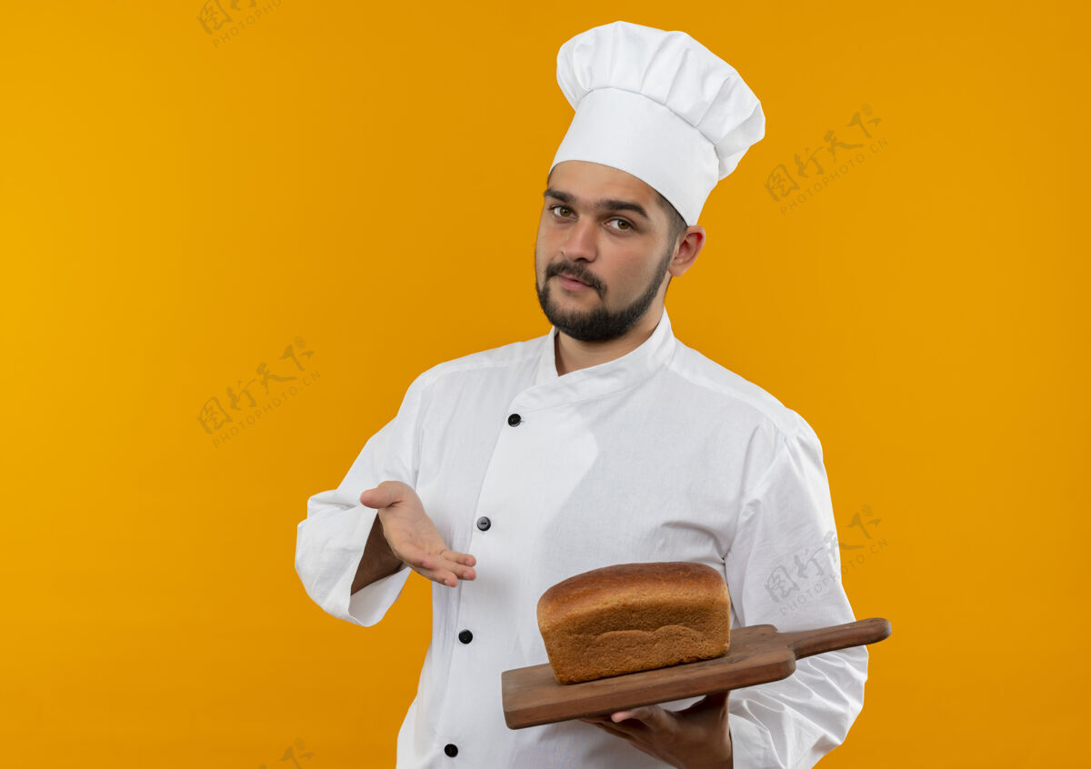 烹饪身着厨师制服的自信的年轻男厨师手拿着并指着橘色墙上挂着面包的砧板信心制服切割