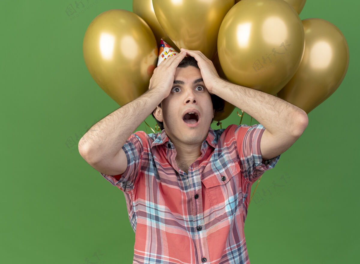 氦焦急英俊的白人男子戴着生日帽站在氦气球前把手放在头上前面空间生日