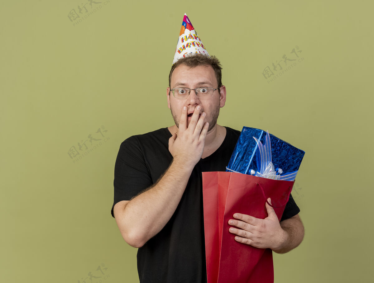 生日震惊的成年斯拉夫人戴着眼镜戴着生日帽 手放在嘴上 手里拿着购物袋里的礼品盒眼镜穿购物